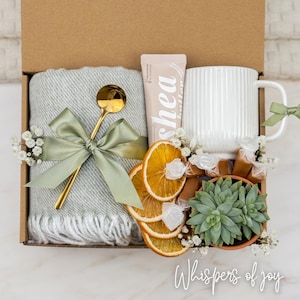 May include: A gift box with a gray blanket, gold spoon, white mug, and a tube of shea lotion. The box also contains dried orange slices, a small succulent, and wrapped candies. The box is tied with a green ribbon and has the text "Whispers of joy".