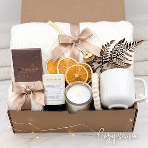 May include: A gift box filled with cozy items. It includes a white blanket, a chocolate bar, a candle, a mug, dried orange slices, and a gold spoon. The box is tied with a satin ribbon. The text "Cozy season" is written on the bottom right.