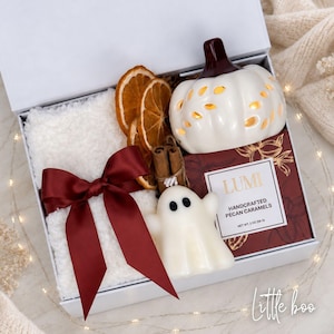 Op de afbeelding: Een witte geschenkdoos met een witte pluizige deken vastgebonden met een donkerrood satijnen lint, twee gedroogde sinaasappelschijfjes, twee kaneelstokjes, een witte keramische pompoenlantaarn, een wit spookfiguurtje en een doosje pecannootkaramels.