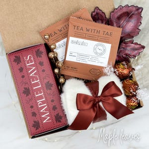 May include: A gift box with a fall theme, featuring a box of "Maple Leaves" tea, two "Tea With Tae" tea packets, a white fluffy item with a burgundy bow, and chocolate candies. The box is decorated with burgundy maple leaf accents.