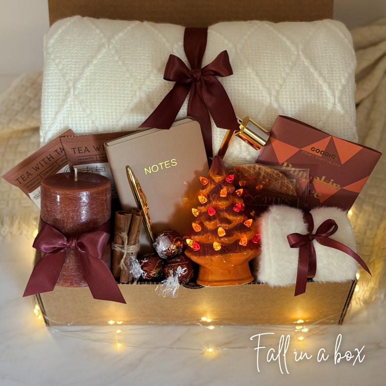 May include: A gift box filled with fall-themed items. Includes a cream-colored blanket, a candle with a burgundy bow, a notebook, a pen, a ceramic tree with lights, chocolates, tea packets, and a fluffy white item with a burgundy bow. The box has the text "Fall in a box".