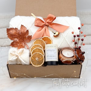 May include: A gift box filled with a white cable-knit blanket, dried orange slices, a candle, and a tea container. The box is decorated with a copper-colored leaf, a bow, and a decorative spoon. The text "Fall into comfort" is at the bottom.