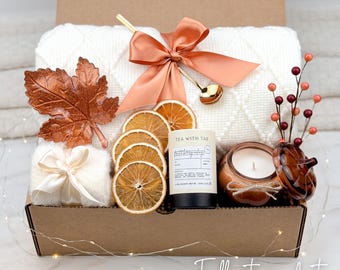 May include: A gift box containing a white knit blanket with a copper satin bow, a copper-colored leaf ornament, a small white gift box with a cream ribbon, dried orange slices, a Tea With Tae tea tin, a candle, and a decorative berry sprig.
