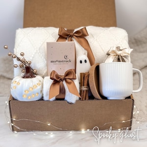 May include: A gift box filled with a white blanket, a white pumpkin with cut-out lights, a white mug, a small ghost figurine, and a box of "moonstruck" products. The box is decorated with brown bows and cinnamon sticks.