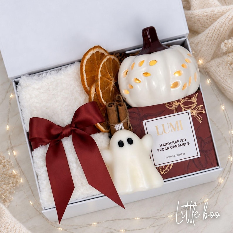 May include: A white gift box containing a white fuzzy blanket tied with a dark red satin ribbon, two dried orange slices, two cinnamon sticks, a white ceramic pumpkin lantern, a white ghost figurine, and a box of Lumi Handcrafted Pecan Caramels.