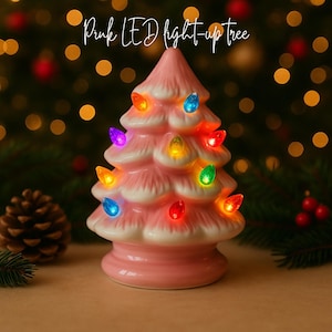 May include: A pink ceramic Christmas tree with white accents and colorful LED lights. The tree is shaped like a traditional fir tree and has a glossy finish. The text "Pink LED light-up tree" is written in cursive at the top.