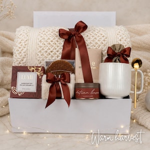 Op de afbeelding: Een witte geschenkdoos met een cr&egrave;mekleurige kabelgebreide deken vastgebonden met een donkerbruin satijnen lint, een doos Lumi handgemaakte pecannootkaramels, een stroopwafel, een geurkaars met het label Autumn Leaves, een witte mok en een gouden lepel.