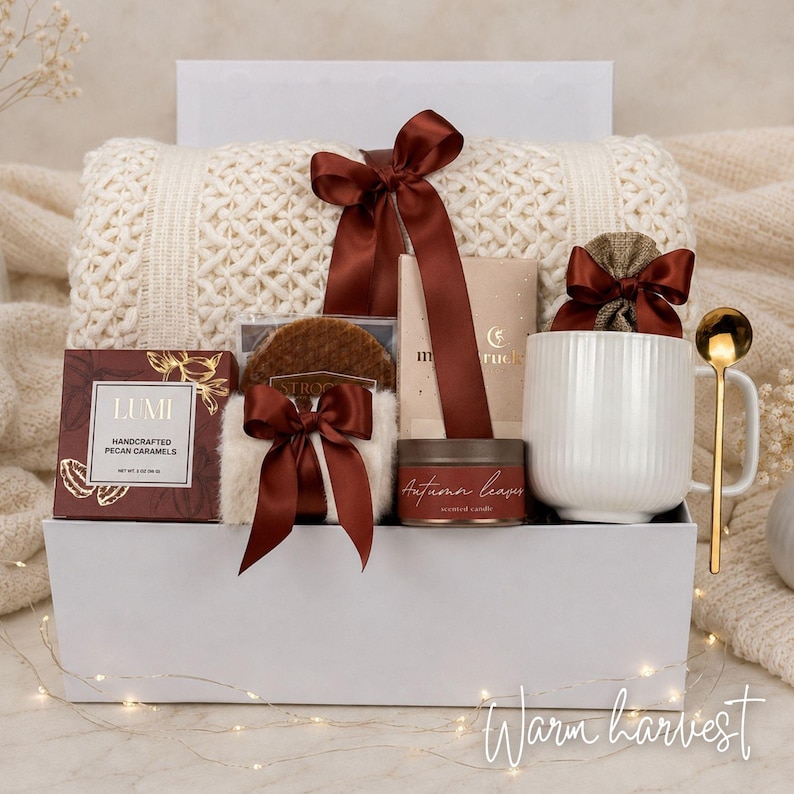 Op de afbeelding: Een witte geschenkdoos met een cr&egrave;mekleurige kabelgebreide deken vastgebonden met een donkerbruin satijnen lint, een doos Lumi handgemaakte pecannootkaramels, een stroopwafel, een geurkaars met het label Autumn Leaves, een witte mok en een gouden lepel.