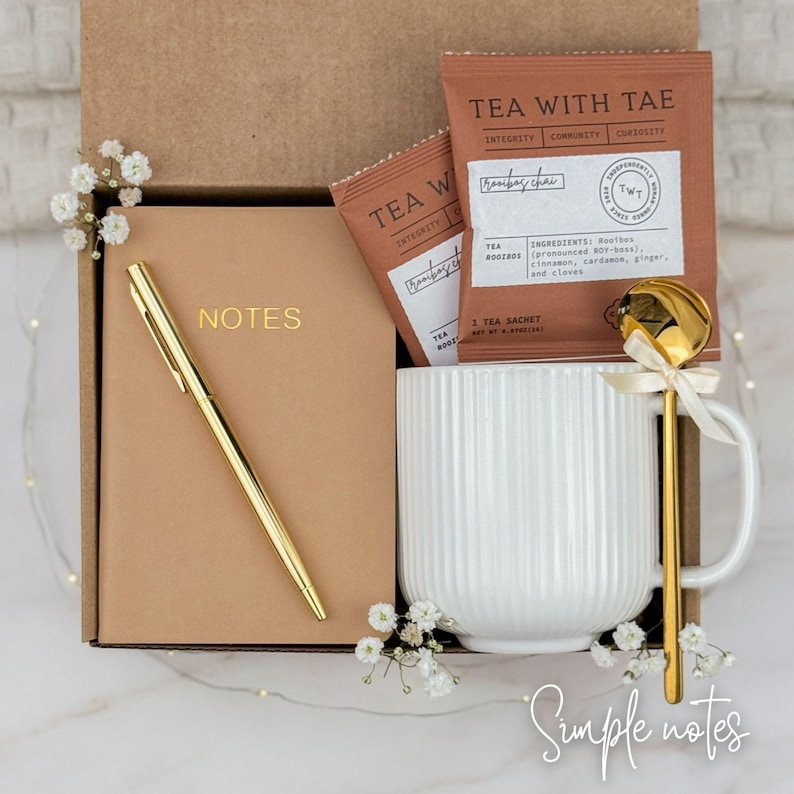 May include: A gift box containing a tan notebook with the word NOTES in gold, a gold pen, a white ribbed ceramic mug, a gold spoon with a small ribbon, and two tea sachets labeled TEA WITH TAE Rooibos Chai, all accented with small white flowers.