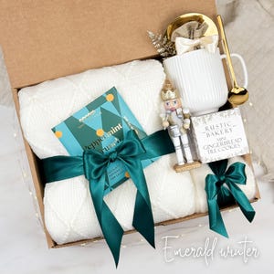 May include: A gift box with a white knitted blanket, a mug, a gold spoon, a nutcracker, a chocolate bar, and a box of cookies. The blanket is tied with a dark green ribbon. The chocolate bar says "Peppermint Crunch". The box is labeled "Emerald winter".
