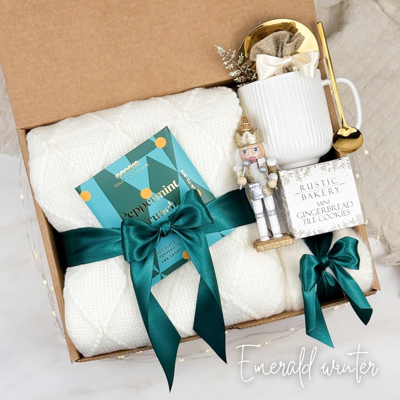 May include: A gift box with a white knitted blanket, a mug, a gold spoon, a nutcracker, a chocolate bar, and a box of cookies. The blanket is tied with a dark green ribbon. The chocolate bar says "Peppermint Crunch". The box is labeled "Emerald winter".