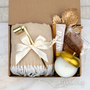 May include: A gift box filled with a beige blanket tied with a cream ribbon, a gold candle snuffer, a shea butter tube, a stroopwafel, a gold acorn-shaped candle, and gold leaf accents. The box is labeled "Warm hug".