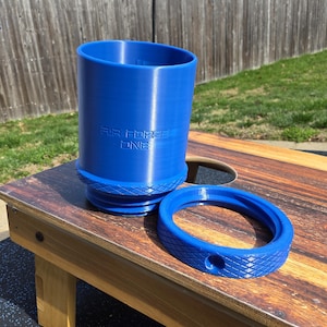 May include: A blue cylindrical container with the text "AIR FORCE ONE" embossed on the side. A matching blue ring sits next to the container. The objects are on a wooden surface.
