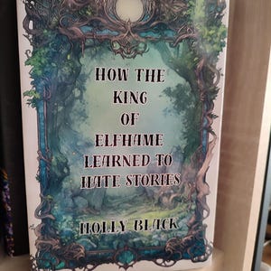 May include: A hardcover book titled "HOW THE KING OF ELFHAME LEARNED TO HATE STORIES" by Holly Black. The cover features a fantasy illustration with a forest scene in shades of blue, green, and white, framed by ornate, dark green and blue foliage.
