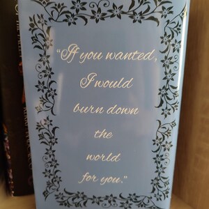 Pu&ograve; includere: Un libro azzurro con la citazione "If you wanted, I would burn down the world for you." in scrittura bianca. Il libro ha un bordo floreale nero ed &egrave; esposto su una mensola di legno.