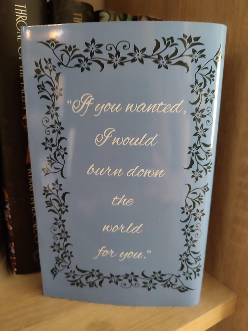 Pu&ograve; includere: Un libro azzurro con la citazione "If you wanted, I would burn down the world for you." in scrittura bianca. Il libro ha un bordo floreale nero ed &egrave; esposto su una mensola di legno.