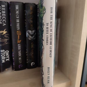 May include: A book titled "How the King of Elfhame Learned to Hate Stories" by Holly Black. The book's spine is white with green and blue illustrations. Other books with dark covers are visible on a wooden shelf.