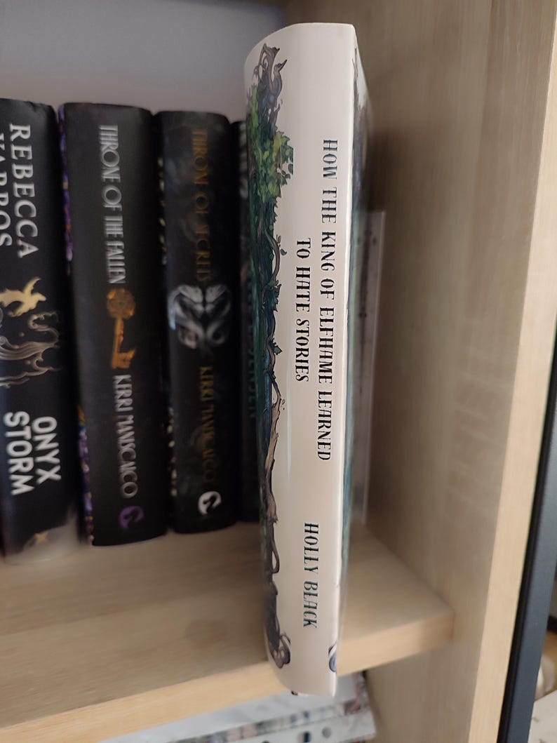 May include: A book titled "How the King of Elfhame Learned to Hate Stories" by Holly Black. The book's spine is white with green and blue illustrations. Other books with dark covers are visible on a wooden shelf.