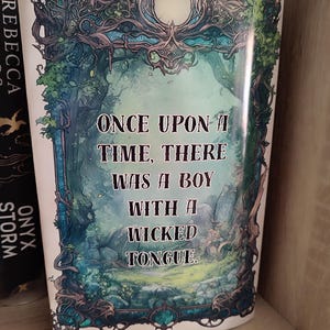 May include: A book cover with the text "ONCE UPON A TIME, THERE WAS A BOY WITH A WICKED TONGUE." The cover features a fantasy illustration with a forest scene in shades of green and blue, framed by ornate, vine-like details. The book is on a shelf.