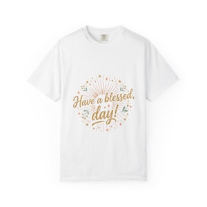 Könnte beinhalten: Weißes T-Shirt mit einem goldenen Design. Das Design zeigt den Text "Have a blessed, day!" in einer Schriftart, umgeben von einem Sonnenaufgang, Sternen und floralen Elementen.