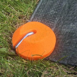 May include: A bright orange circular tent peg anchor with a smooth, flat top surface and a central metallic hook. The anchor is positioned to secure the edge of a dark grey, woven textured tarp, providing a stable point for outdoor equipment.