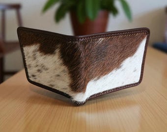 May include: A bi-fold wallet featuring a brown and white cowhide pattern. The wallet has a dark brown leather trim and stitching. The wallet is made of genuine leather and is designed to hold cards and cash.