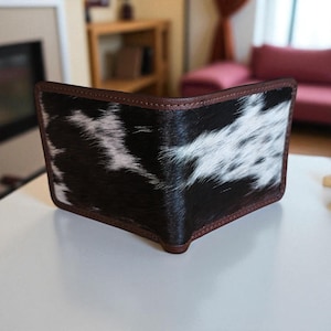 Puede incluir: Una cartera plegable con un patrón de piel de vaca en blanco y negro, enmarcada por un ribete de cuero marrón. La cartera está abierta, mostrando el diseño interior. El producto está diseñado para llevar dinero en efectivo y tarjetas.