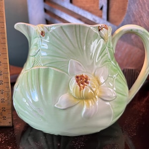 May include: A light green ceramic jug with a white and yellow flower design. The jug has a curved handle and a spout for pouring. A wooden ruler is next to the jug, showing measurements in inches.