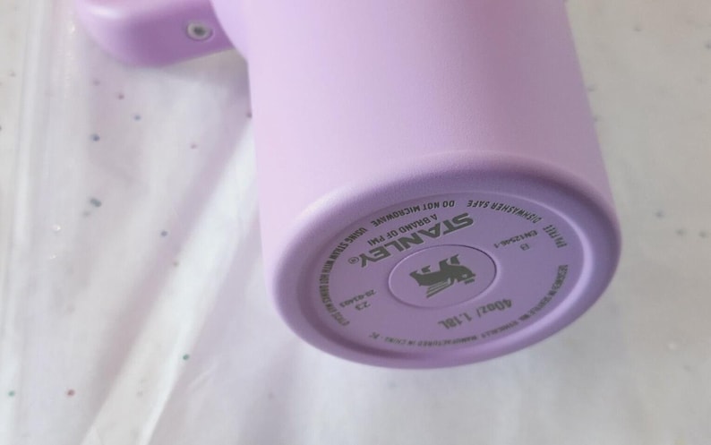 Puede incluir: Un vaso Stanley de color lila claro. La parte inferior del vaso tiene el nombre de la marca, el logotipo y otro texto. El vaso es de 1,18 L. El vaso est&aacute; sobre una superficie blanca con peque&ntilde;as motas de colores.