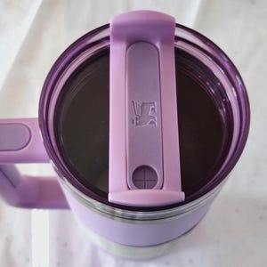 Puede incluir: Un termo de viaje aislado de color lavanda con asa y tapa a juego. La tapa tiene un cierre abatible con un logotipo. El termo tiene un interior de acero inoxidable y una banda blanca en el medio.