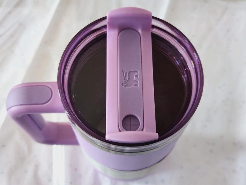 Puede incluir: Un termo de viaje aislado de color lavanda con asa y tapa a juego. La tapa tiene un cierre abatible con un logotipo. El termo tiene un interior de acero inoxidable y una banda blanca en el medio.