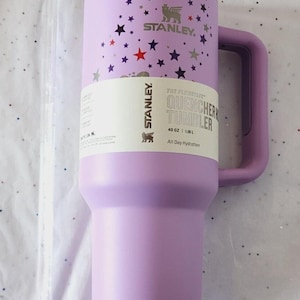 Puede incluir: Un vaso Stanley Quencher lavanda con asa, decorado con estrellas. El vaso tiene una capacidad de 1,18 L y presenta el logotipo de Stanley y el texto "All Day Hydration".