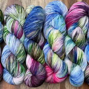 Könnte beinhalten: Sechs handgefärbte Garnstränge in einer Reihe angeordnet, jeder mit einer bunten Farbpalette aus leuchtendem Pink, tiefem Violett, hellem Blau und Limettengrün, vermischt mit Weiß, was ein mehrfarbiges, gesprenkeltes und gestreiftes Aussehen ergibt.