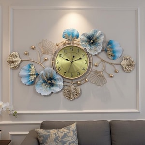 May include: A decorative wall clock with a gold-coloured face and hands, surrounded by blue and gold floral accents. The clock is mounted on a white wall, with a sofa and artwork visible below.