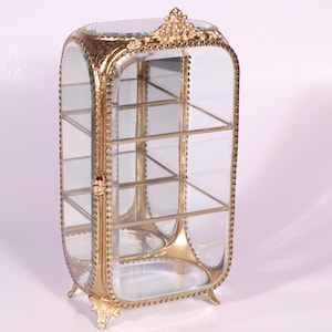 Puede incluir: Una vitrina vertical con un marco de metal dorado con detalles ornamentados, paneles de vidrio transparente en los laterales y el frente, y tres estantes interiores de vidrio. La estructura se apoya sobre cuatro patas decorativas e incluye un pestillo.