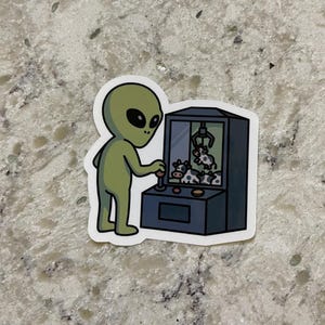 May include: A sticker featuring a green alien operating a claw machine. The claw machine contains small cow plush toys. The alien is depicted with large black eyes and is wearing a green outfit. The sticker has a white border.