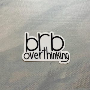 May include: A white sticker with black text that reads "brb overthinking." The sticker has a black border and is on a light blue and gray background. The sticker is rectangular in shape.