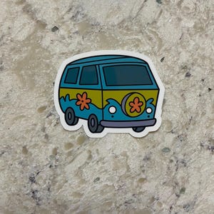 May include: A sticker featuring a cartoon van in shades of blue, yellow, and green. The van has flower designs on the side and a purple bumper. The sticker is on a white background.
