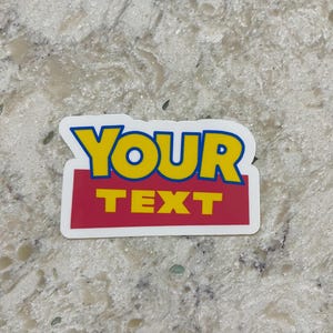 May include: A rectangular sticker with the words "YOUR TEXT" in yellow and red, outlined in blue. The sticker has a white border and is set against a speckled, light-colored surface.