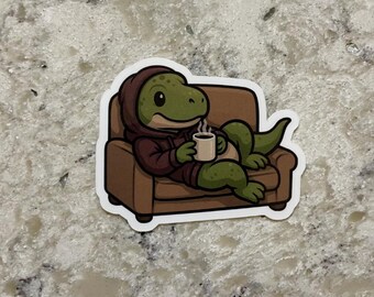 May include: A sticker featuring a cartoon dinosaur in a brown hoodie, relaxing on a brown couch and holding a steaming mug. The dinosaur is green with a light green belly. The sticker has a white border.