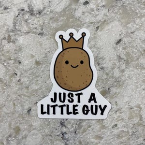 May include: A sticker featuring a cartoon potato wearing a gold crown. The potato has a smiling face and is brown with small speckles. The text "JUST A LITTLE GUY" is below the potato in black letters.