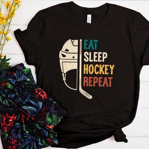 May include: Black t-shirt with a hockey-themed design. The shirt features a hockey helmet and stick graphic, with the words "EAT SLEEP HOCKEY REPEAT" in teal, yellow, and red. The shirt is paired with floral patterned pants and yellow flowers.