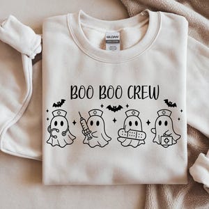 Op de afbeelding: Crèmekleurige sweatshirt met de zwarte tekst "BOO BOO CREW". Onder de tekst staan vier spookkarakters verkleed als verpleegsters, elk met medische hulpmiddelen. Het ontwerp bevat ook vleermuizen en sterren.
