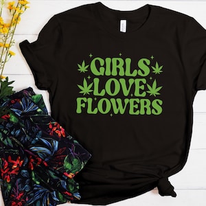 May include: Black t-shirt with the text "GIRLS LOVE FLOWERS" in green, featuring cannabis leaf graphics. The shirt is styled with patterned trousers and a bouquet of yellow flowers.
