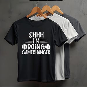 Könnte beinhalten: Drei T-Shirts hängen an einer Holzstange. Die Shirts sind schwarz, weiß und grau. Das schwarze T-Shirt hat den weißen Text "SHHH I'M DOING GAMECHANGER". Die Shirts sind kurzärmlig.