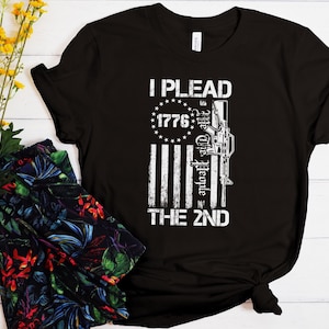 Op de afbeelding: Zwart t-shirt met een verweerde witte afbeelding. Het ontwerp bevat de tekst "I Plead The 2nd" met een gestileerde Amerikaanse vlag en een geweersilhouet. Het jaar "1776" staat in een cirkel van sterren.