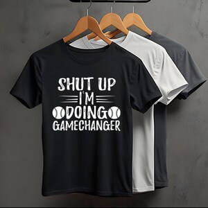 Könnte beinhalten: Drei T-Shirts hängen an Holzbügeln. Das vordere T-Shirt ist schwarz mit weißem Text "SHUT UP I'M DOING GAMECHANGER" mit Baseball-Grafiken. Die anderen beiden T-Shirts sind weiß und grau.
