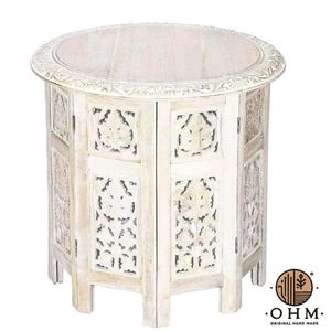 Pode incluir: Uma mesa lateral redonda com acabamento caiado, apresentando padrões florais esculpidos complexos nos painéis laterais e uma borda decorativa ao redor do topo. A mesa apoia-se em várias pernas e tem uma textura de madeira natural clara.