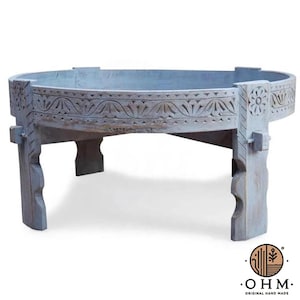 Puede incluir: Una mesa redonda y baja de madera con un acabado azul claro desgastado. La mesa presenta intrincados patrones florales y geométricos tallados a mano en el borde exterior y patas gruesas y resistentes con un diseño de bordes festoneados.