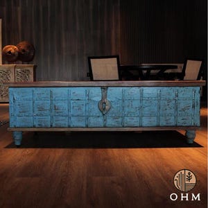 Puede incluir: Un baúl de almacenamiento rectangular de madera con un acabado de pintura azul claro desgastado en el panel frontal, dividido en un patrón de cuadrícula. El baúl tiene una parte superior y base de madera marrón oscuro, con cuatro patas cortas y redondeadas.